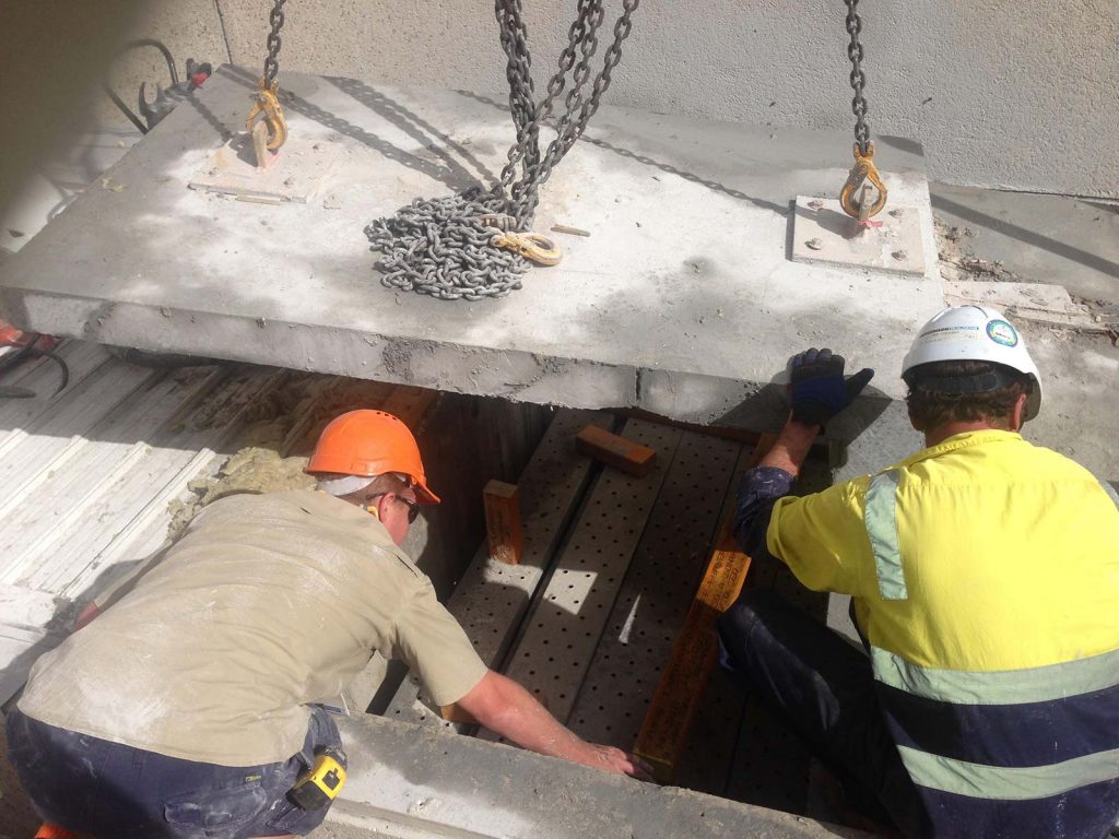 two construction workers wearing protective clothing and hard hats moving a large slab of concrete with metal chains